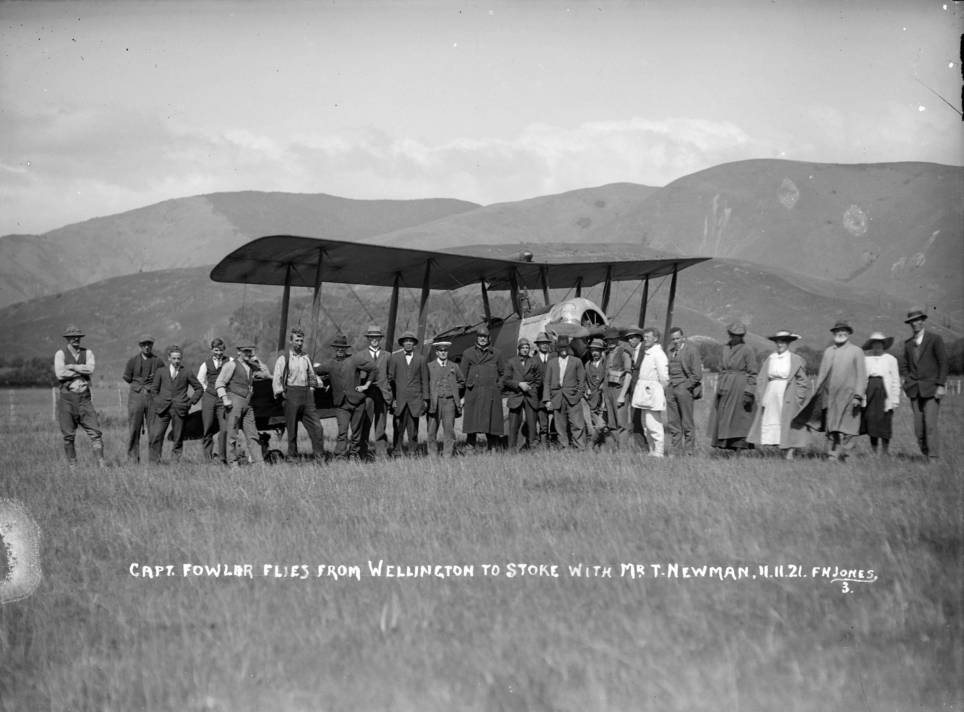 Black and white photo a biplane parked in a field surrounded by a crowd.